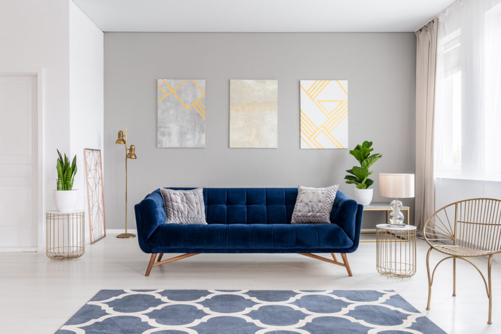 An elegant navy blue sofa in the middle of a bright living room interior with gold metal side tables and three paintings on a gray wall. Real photo.